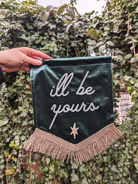 Gold-fringed "i'll be yours" text on green velvet banner against ivy leaf backdrop.