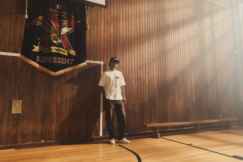 A man dressed in a white t-shirt is located in a wooden sports hall, with a large banner hanging from the netball hoop above.