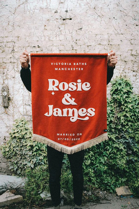  Elegant terracotta velvet wedding banner displaying "Victoria Baths", "Rosie and James", and "07/05/2022" the background is a white brick wall with ivy.