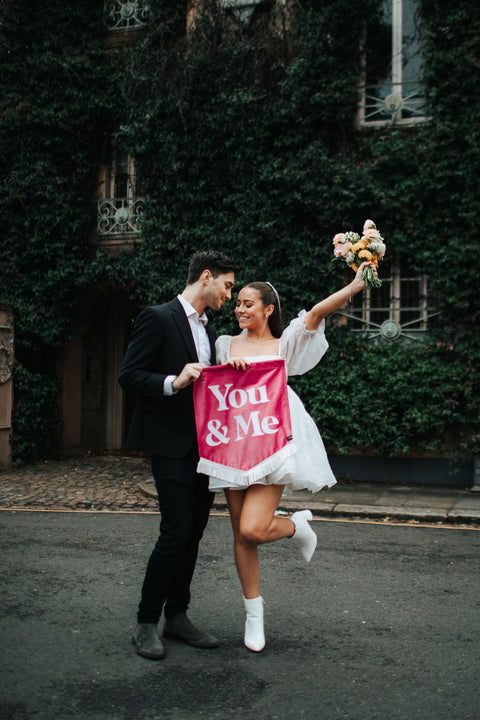 Velvet wedding mini banner in pink with "you and me" text, white fringing, held by married couple