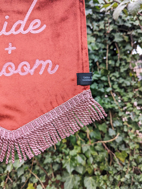 Velvet banner in terracotta with "bride and groom" pink text, and pink fringing on ivy leaf background.