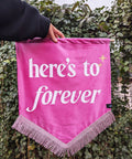 Velvet wedding banner in pink with "heres to forever" white text, pink fringing and gold star applique, on ivy leaf background.