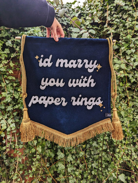 Velvet wedding banner in navy with "id marry you with paper rings" text, gold fringing and hanging tassels, on ivy leaf background.