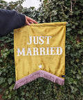 Velvet wedding banner in yellow with "just married" white text and pink fringing on ivy leaf background.