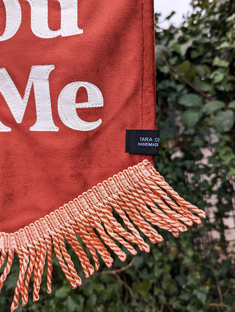 Velvet wedding banner in terracotta with "You and Me" text and peach fringing on ivy leaf background.