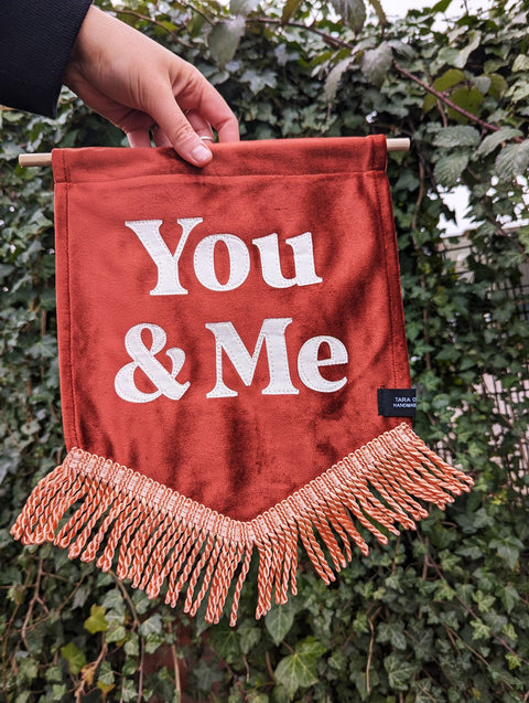 Velvet wedding banner in terracotta with "You and Me" text and peach fringing on ivy leaf background.