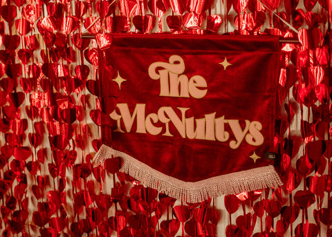 Deep red wedding banner reading "The McNultys," adorned with metallic love heart decorations on a wall.