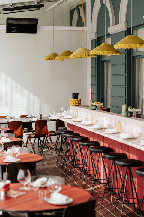 Contemporary Italian restaurant interior featuring low bar, bar stools, tables set with wine glasses and cutlery. Yellow lampshades adorn the ceiling.