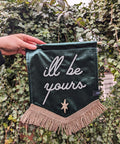 Gold-fringed "i'll be yours" text on green velvet banner against ivy leaf backdrop.