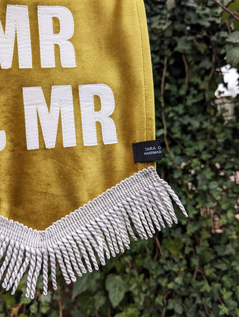 Velvet wedding banner in gold with "mr and mr" white text and white fringing detail on ivy leaf background.