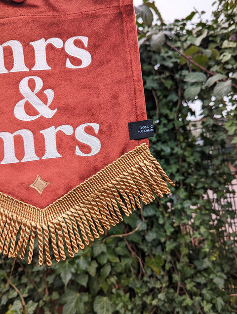Velvet mini banner in terracotta with "mrs & mrs" text, gold fringing and gold star applique on ivy leaf background.