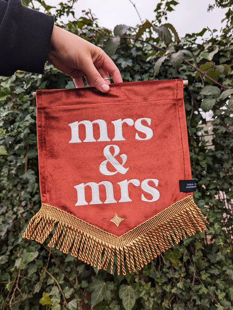 Velvet mini banner in terracotta with "mrs & mrs" text and gold fringing on ivy leaf background.