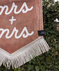 Velvet wedding banner in pink with "mrs and mrs" white text and white fringing detail on ivy leaf background.