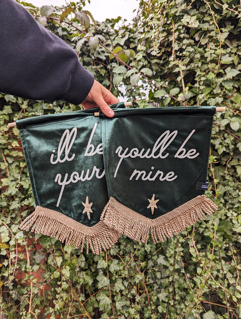 Two green velvet banners with gold fringing, one saying "I'll be yours" and the other "You'll be mine", against an ivy backdrop.