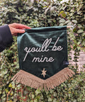 Gold-fringed "you'll be mine" text on green velvet banner against ivy leaf backdrop.