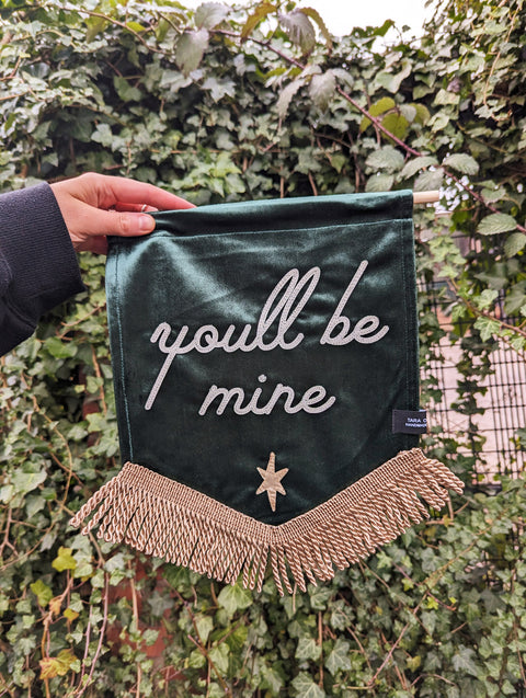 Gold-fringed "you'll be mine" text on green velvet banner against ivy leaf backdrop.