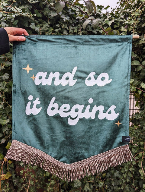 Velvet banner in green with "and so it begins" text, with bronze fringing and gold star appliques on ivy leaf background.