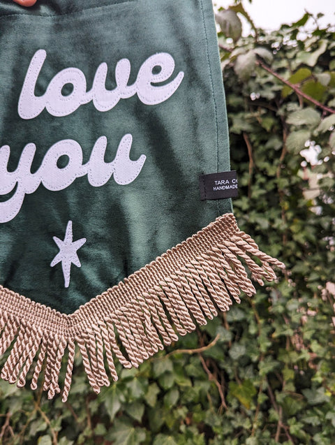 Ivy leaf backdrop with close up of green velvet banner, ivory gold fringing, and "i love you" text.