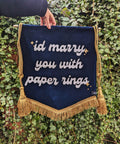 Velvet wedding banner in navy with "id marry you with paper rings" text, gold fringing and hanging tassels, on ivy leaf background.