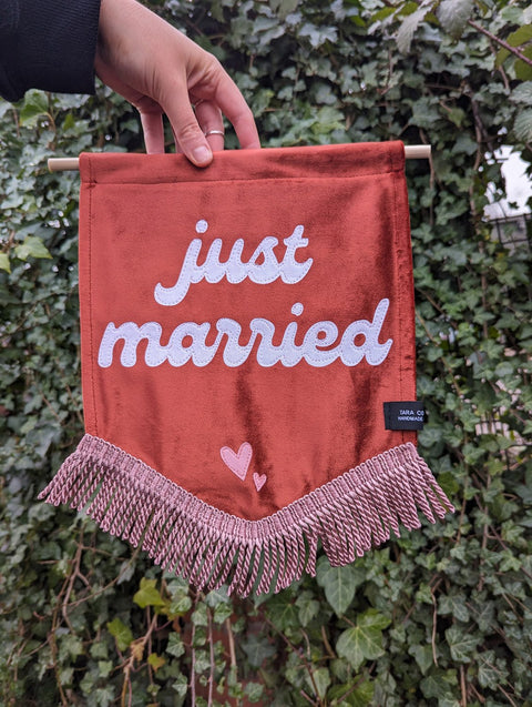 Velvet banner in terracotta with "just married" text with pink fringing on ivy leaf background.