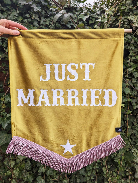Velvet wedding banner in yellow with "just married" white text and pink fringing on ivy leaf background.