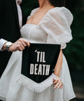 Close up of velvet wedding mini banner in black with "til death" text and white fringing held by married couple.