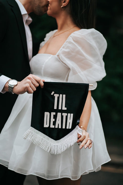 Close up of velvet wedding mini banner in black with "til death" text and white fringing held by married couple.
